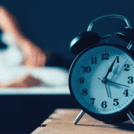 Alarm clock on bedside table in front of a blurred out image of a person in bed